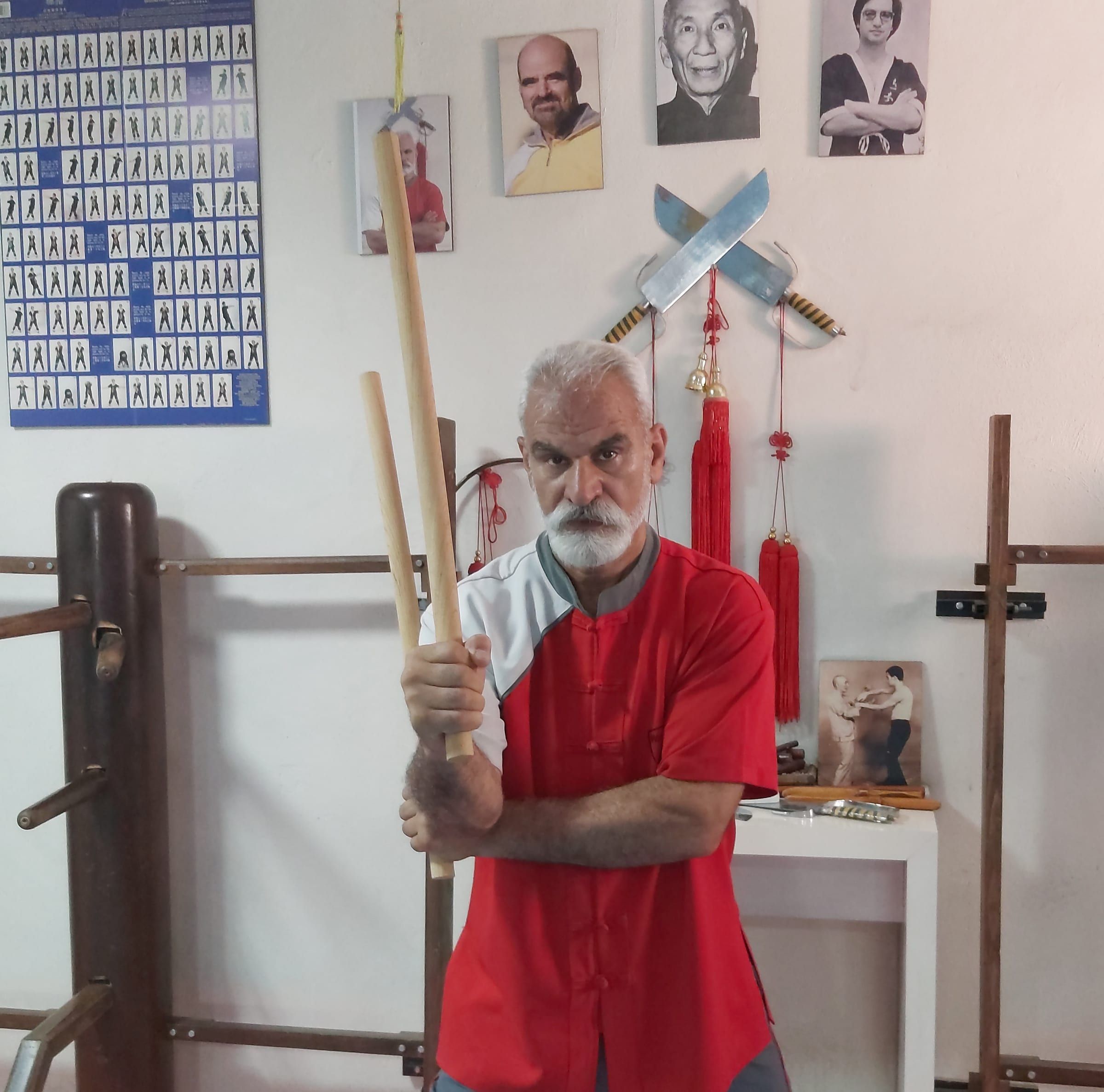Wing-Tsun-Lehrer mit Escrima-Stöcken in einem Trainingsraum mit Holzpuppe und Doppelmessern an der Wand